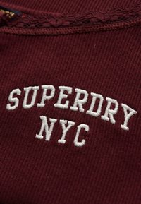 Bordeauxrode geribbelde stof met witte geborduurde tekst "SUPERDRY NYC" en een kanten rand bij de halslijn zichtbaar aan de bovenkant.