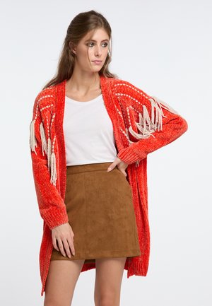 Frau trägt ein weißes Hemd, einen braunen Rock und eine rote Strickjacke mit weißen Fransen, posiert mit der Hand in der Hüfte.