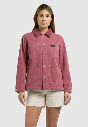 Naine, kes kannab nupuga kinni pandud tolmuroosa jopet, millel on neli esist taskut, ja kreemikaid lühikesi pükse, seisab hallil ühevärvilisel taustal.