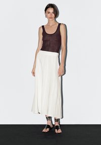 Model stands against a plain white background, wearing a dark tank top, white flowing skirt, and stylish black sandals, with loose hair.