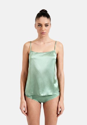 Woman with hair in a bun wearing a light green satin camisole and matching underwear, standing against a plain light background.
