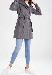 Šedý kabát s kapucí a zavazováním v pase, se strukturovaným designem a měkkou texturou. Kombinován s tmavě modrými úzkými džíny a bílými teniskami.