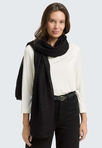 Sciarpa nera a coste con sottili scintillii, drappeggiata attorno al collo. Indossata con una maglietta bianca e pantaloni neri, completati da una cintura con fibbia dorata.