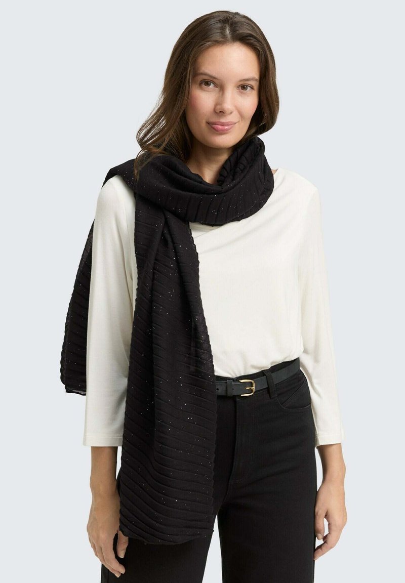 Sciarpa nera a coste con sottili scintillii, drappeggiata attorno al collo. Indossata con una maglietta bianca e pantaloni neri, completati da una cintura con fibbia dorata.