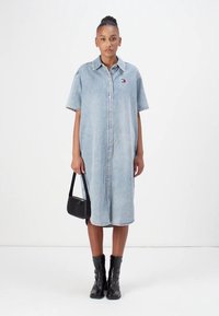 Jeansklänning, ljusblå, kortärmad, med krage, knappar ned fram, fransad nederkant. Modellen håller en liten svart handväska och bär svarta stövlar.
