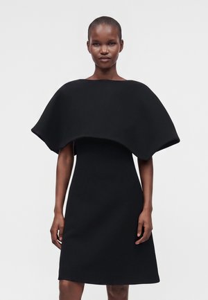 Femme portant une robe noire avec une large surcouche structurée en forme de cape, debout devant un fond clair uni.