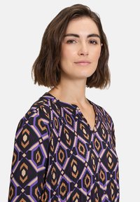 Black blouse featuring a geometric pattern in shades of orange, purple, and cream. It has a V-neckline and relaxed sleeves with pleats.