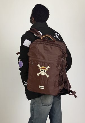 Mochila marrón con un gráfico de calavera y huesos cruzados. Cuenta con correas ajustables, bolsillos con cremallera y un exterior texturizado.