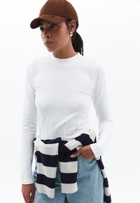 Weißes Langarmshirt mit hohem Kragen; navy- und weiß gestreifter Pullover um die Taille gebunden; braune Kappe. Jeanshose mit lockerer Passform.