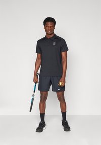 Black polo shirt with a front logo, paired with dark shorts. Holding a tennis racket and a yellow tennis ball, wearing black athletic shoes.