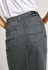Grijze denimrok met een getailleerde pasvorm, voorzien van twee achterzakken, contrasterende stiksels en een zwart leren merklabel aan de taille.