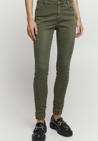Chinos verdes de algodón elástico con cinco bolsillos, ajuste tapered y textura arrugada, combinados con mocasines negros de chunky con herrajes dorados.