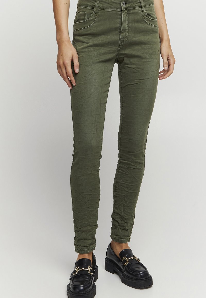 Chinos verdes de algodón elástico con cinco bolsillos, ajuste tapered y textura arrugada, combinados con mocasines negros de chunky con herrajes dorados.
