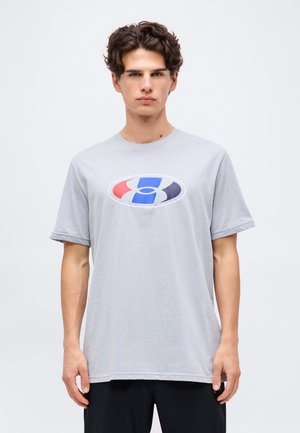 Jonge man draagt een lichtgrijs t-shirt met een rood, blauw en zwart ovaal afbeelding, staand tegen een effen witte achtergrond.