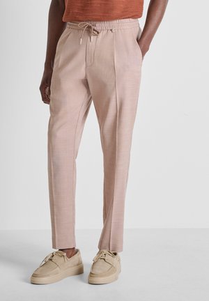 Persona con pantalones de cordón rosa claro y zapatos beige con cordones, de pie con una mano en el bolsillo contra un fondo liso.