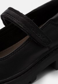 Zapato de cuero negro con una correa de velcro, superficie texturizada, interior acolchado y suela de goma gruesa. Presenta un sutil logotipo en la correa.