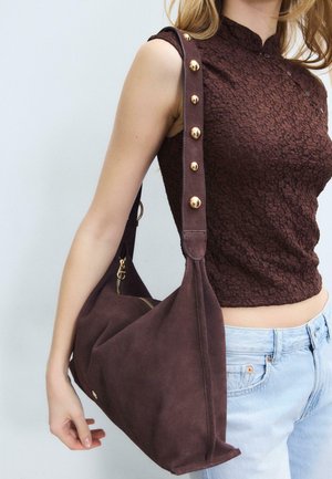 Donna che indossa una maglietta senza maniche di colore marrone scuro e jeans azzurri, che tiene una grande borsa a tracolla in camoscio marrone con dettagli dorati sul cinturino.