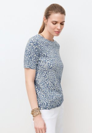 Vaaleanruskeat hiukset poninhännällä, sininen kuviollinen lyhythihainen paita, valkoiset housut ja kultainen ketjukoru, yksivärinen tausta.