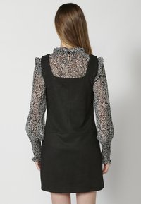 Robe en daim noire avec un décolleté carré, superposée à un haut transparent à motif floral noir et blanc avec des manches à volants.