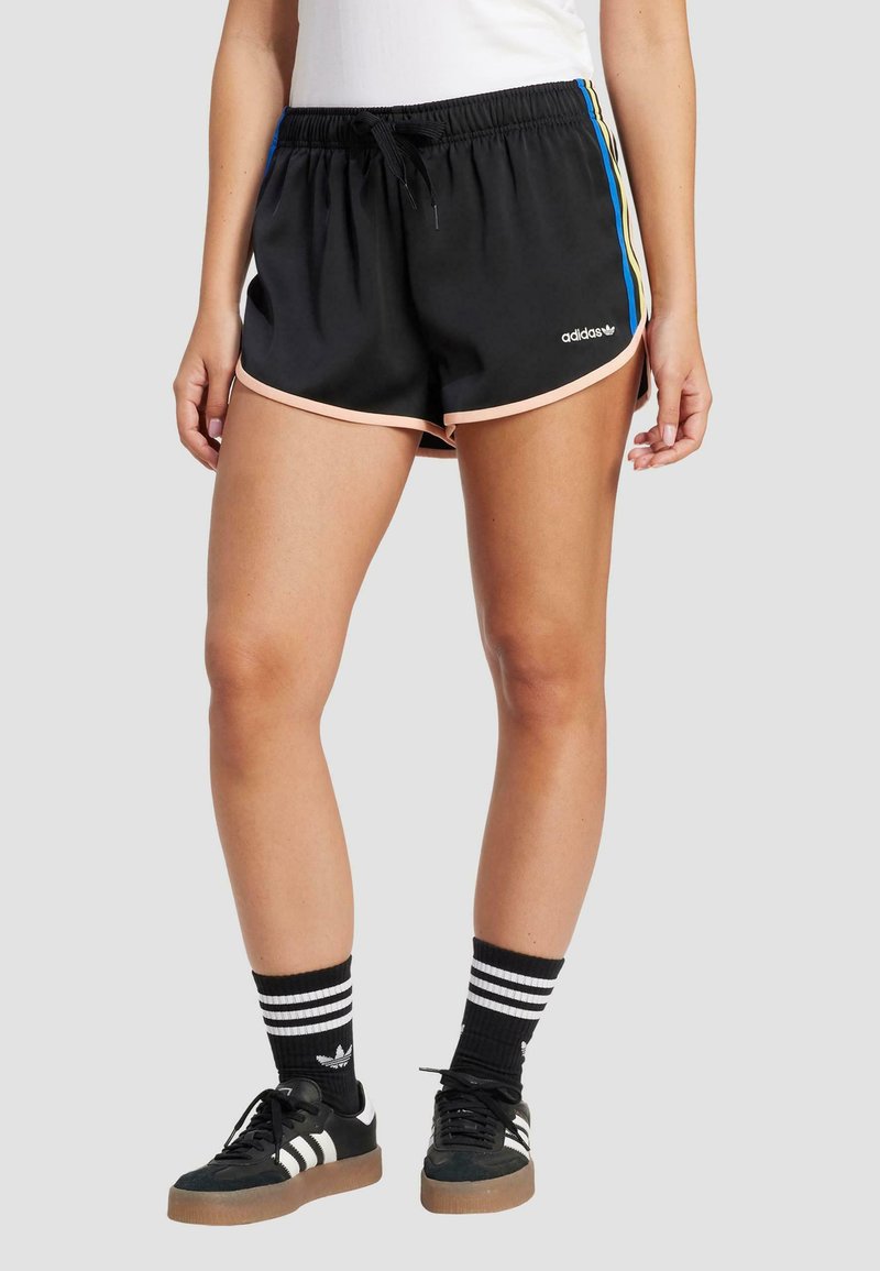 Schwarze Sportshorts mit pinkem Besatz, elastischem Bund und seitlichen Streifen in Blau, Gelb und Weiß. Kombiniert mit schwarzen Turnschuhen und gestreiften Socken.
