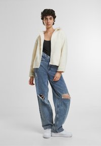 Fleece zip-up jakke i off-white, parret med slidte blå jeans. Har ribbede manchetter og en klassisk pasform, båret med hvide sneakers.