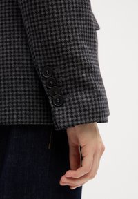 Blazer de espiga gris con una tela texturizada, que presenta cuatro botones negros en el puño y una forma estructurada y ajustada.