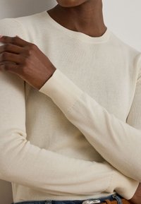 Uomo che indossa un maglione a maglia beige a maniche lunghe con polsini a coste e colletto girocollo, con le braccia incrociate sul petto.