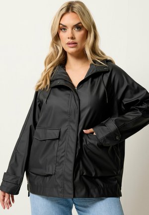 Blonde vrouw die een zwarte waterdichte jas met ritssluiting, klepzakken en manchetten draagt, gecombineerd met lichtblauwe spijkerbroek, staand tegen een effen achtergrond.