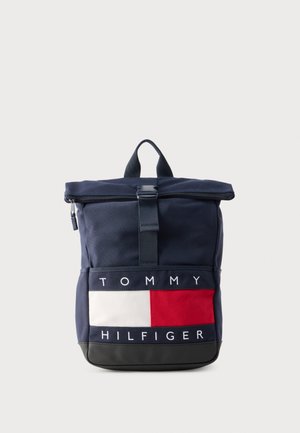 Mørkeblå rygsæk med rulle-top design, forsynet med en stor hvid, rød og blå logopatch foran og sort bund. Tekstureret stof med robust hardware.