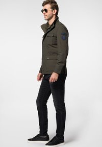 Zielona oliwkowa kurtka z kołnierzem stojącym, kieszeniami z przodu i granatową naszywką; zestawiona z czarnymi dżinsami o slim fit i czarnymi trampkami.