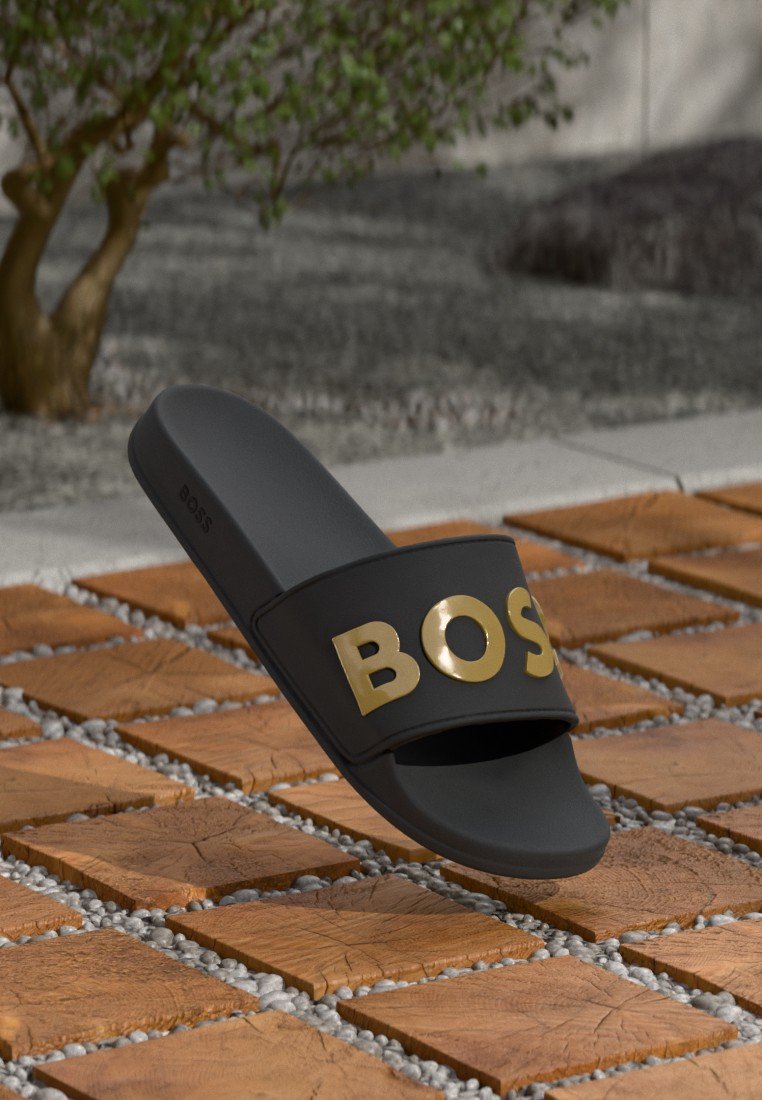 Sandale noire à glissière avec une large sangle arborant un logo "BOSS" en or. La semelle est lisse et légèrement conique, avec une finition mate.