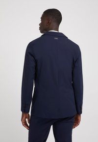Blazer bleumarin cu un croi ajustat, vent unic în spate și etichetă de marcă. Dispune de mâneci lungi și manșete cu butoane.