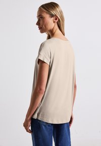 Beige krátký rukáv top s kulatým výstřihem, hladkou texturou a bočními rozparky. V kombinaci s modrými džíny, foceno z bočního úhlu.