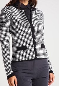 Kvinna iklädd en svart och vit houndstooth-kardigan med svarta kanter och fickor över en svart skjorta med krage, i kombination med mörka byxor.