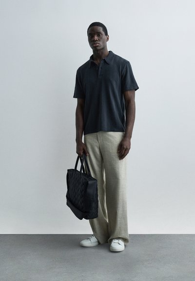 Άνδρας με μαύρο πόλο πουκάμισο, μπεζ παντελόνι φαρδύ στα πόδια, λευκά αθλητικά παπούτσια, κρατώντας μια μαύρη τσάντα tote με καπιτονέ σχέδιο, στέκεται μπροστά σε έναν απλό τοίχο.