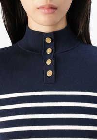 Navy strikket topp med høy krage, med fire gullknapper og horisontale hvite striper på brystet. Glatt tekstur.