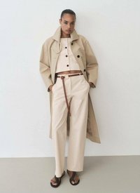Beige trenchcoat över en ljusbeige kort topp och vida byxor, accentuerat med ett brunt bälte. Modellen bär svarta sandaler.