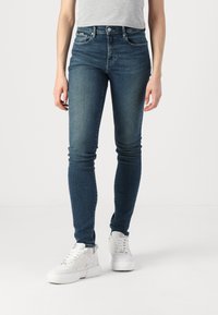 Jeans skinny azul oscuro de mezclilla, con cinco bolsillos, cremallera y una leve textura deslavada, combinados con zapatillas blancas.