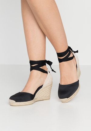 Jambes portant des espadrilles compensées noires avec des talons beiges tissés et des rubans noirs autour de la cheville noués en nœuds.
