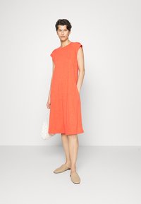 Vestido midi vermelho-alaranjado com mangas curtas, bolsos laterais e um corte descontraído, combinado com sapatilhas bejes e uma bolsa branca trançada.