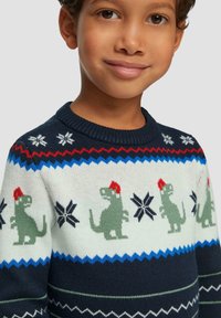 Sweter ma granatowo-kremową kolorystykę z zielonymi wzorami dinozaurów w czerwonych czapkach, płatkami śniegu i zygzakowymi akcentami.