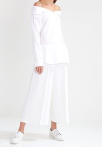Model i en off-the-shoulder hvid skjorte og brede hvide bukser, iført hvide sneakers, poserer mod en ensfarvet baggrund.