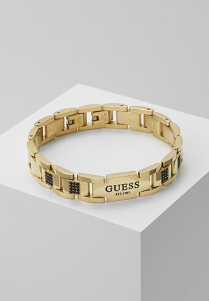 Bracelet en métal doré avec des accents en pierre noire et une plaque gravée de "GUESS EST. 1981" exposée sur un piédestal blanc.