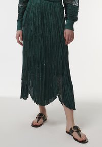 Vestido verde plisado con tejido texturizado, dobladillo asimétrico y acentos dorados. Combinado con sandalias negras con una hebilla decorativa.