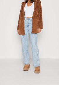 Braune Jacke mit Kapuze aus Kunstfell, weißes Trägershirt, hellblaue, hoch taillierte Jeans und beige Plateauschuhe. Das Outfit hat ein Design mit offenem Vorderteil.