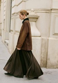 Kvinde med hår i en bun der går ved siden af en stenbygning, iført en brun jakke over en lang, flydende mørk nederdel og mørke sko.