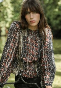 Femme aux longs cheveux bruns portant un blouse à motif floral noir avec des manches bouffantes, se tenant à l'extérieur avec un arrière-plan de verdure flou.