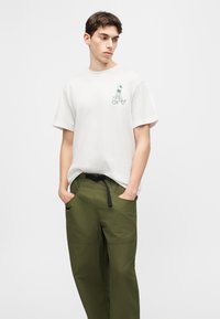 Νέος άνδρας που φοράει λευκό T-shirt με μικρό πράσινο γραφικό αστροναύτη και ελιάς πράσινο παντελόνι με τα χέρια στις τσέπες.