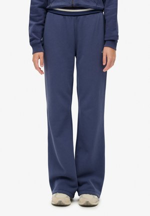 Personne portant un pantalon large bleu marine, une veste assortie et des baskets beige, debout devant un fond blanc uni.