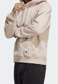 Beige huppari, jossa on kiristettävä huppu, ribbikujat ja etutasku. Taskussa on Adidas-logotunnus. Pehmeä kangaslaatu.